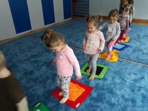 dzieci aktywnie uczestnicząw zajęciach. kilkoro dzieci idzie po ścieżce sensorycznej. Ścieżka to kolorowe kwadraty położone na podłodze